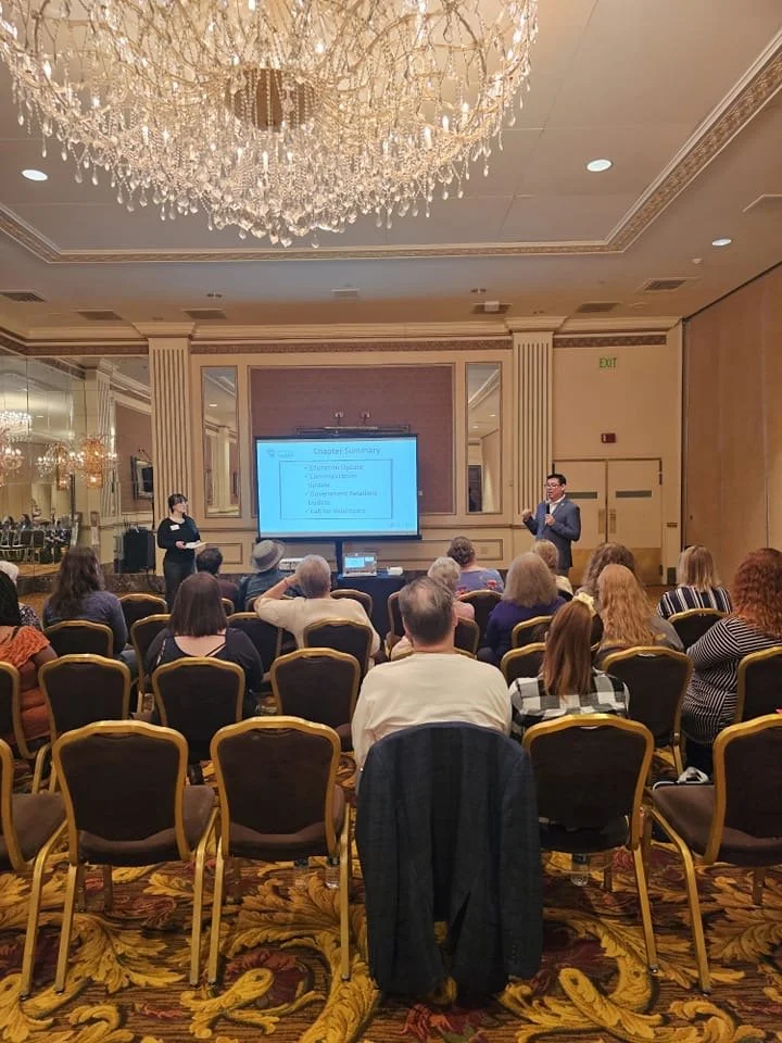 A conference room with a chandelier hanging from the ceiling, where a presentation is in progress. Two speakers stand at the front, one on the left and one on the right, with a large screen displaying slides. The audience is seated in rows of chairs 
