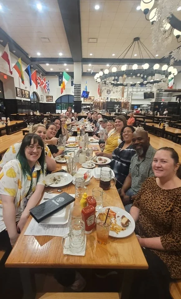 A large group of people sitting around a long wooden table in a restaurant, smiling and enjoying food and drinks, with flags hanging on the walls and large circular chandeliers overhead.