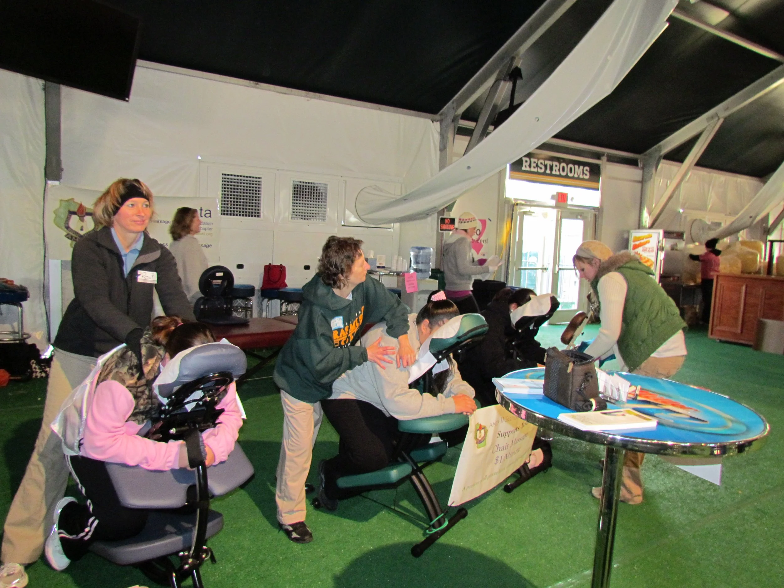 Children and adults gathered around a table inside a large indoor venue, with a sign indicating restrooms and various notices and signs on the wall.