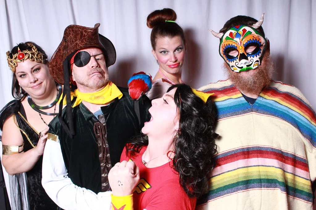 Group of five people dressed in costumes, with one woman in a pirate costume, one man with an eye patch and pirate hat, one woman with a colorful mask and horns, and another woman in a red shirt with a superhero stance, standing in front of a white c