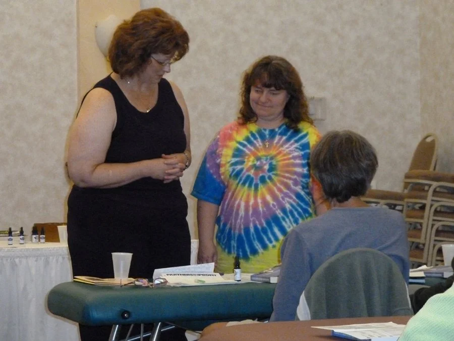 Two women standing and engaging in conversation with a seated man at a table in a room with a beige wall. One woman wears a black sleeveless top, and the other wears a colorful tie-dye shirt. The table has various items, including a small bottle, pap
