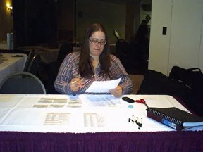 A young woman with glasses sitting at a table, reading or reviewing papers. The table has a purple tablecloth, scissors, papers, and a binder. The background shows a room with chairs and other people.