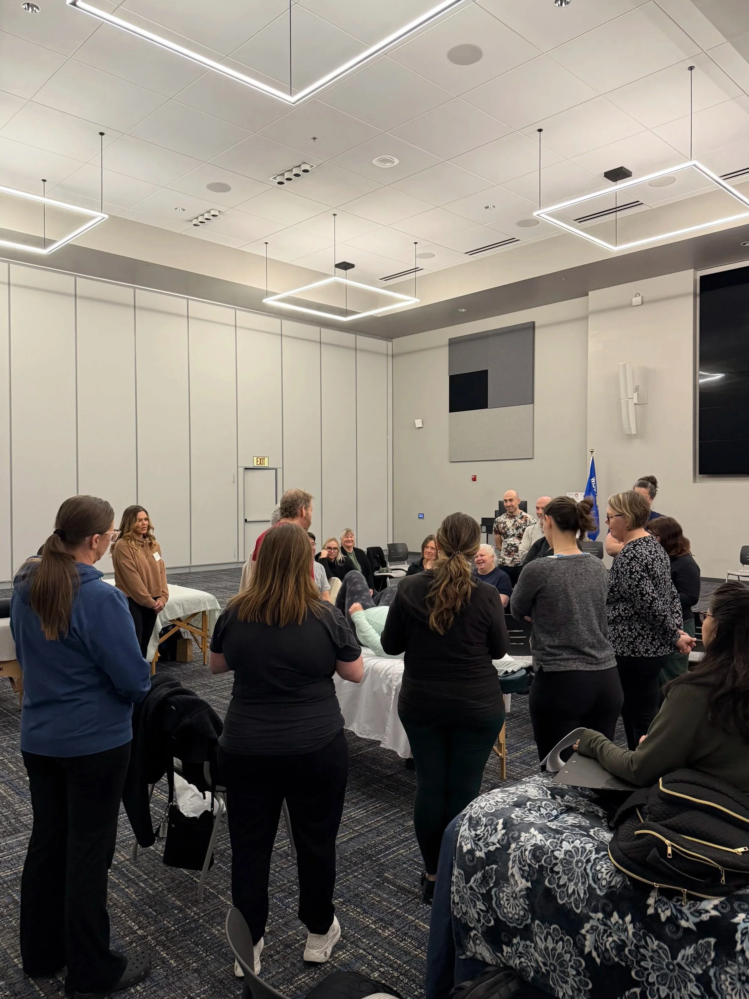 People gathered in a conference room for a group event, some standing around a table with items on it, others sitting and engaging in discussion and listening to a speaker.