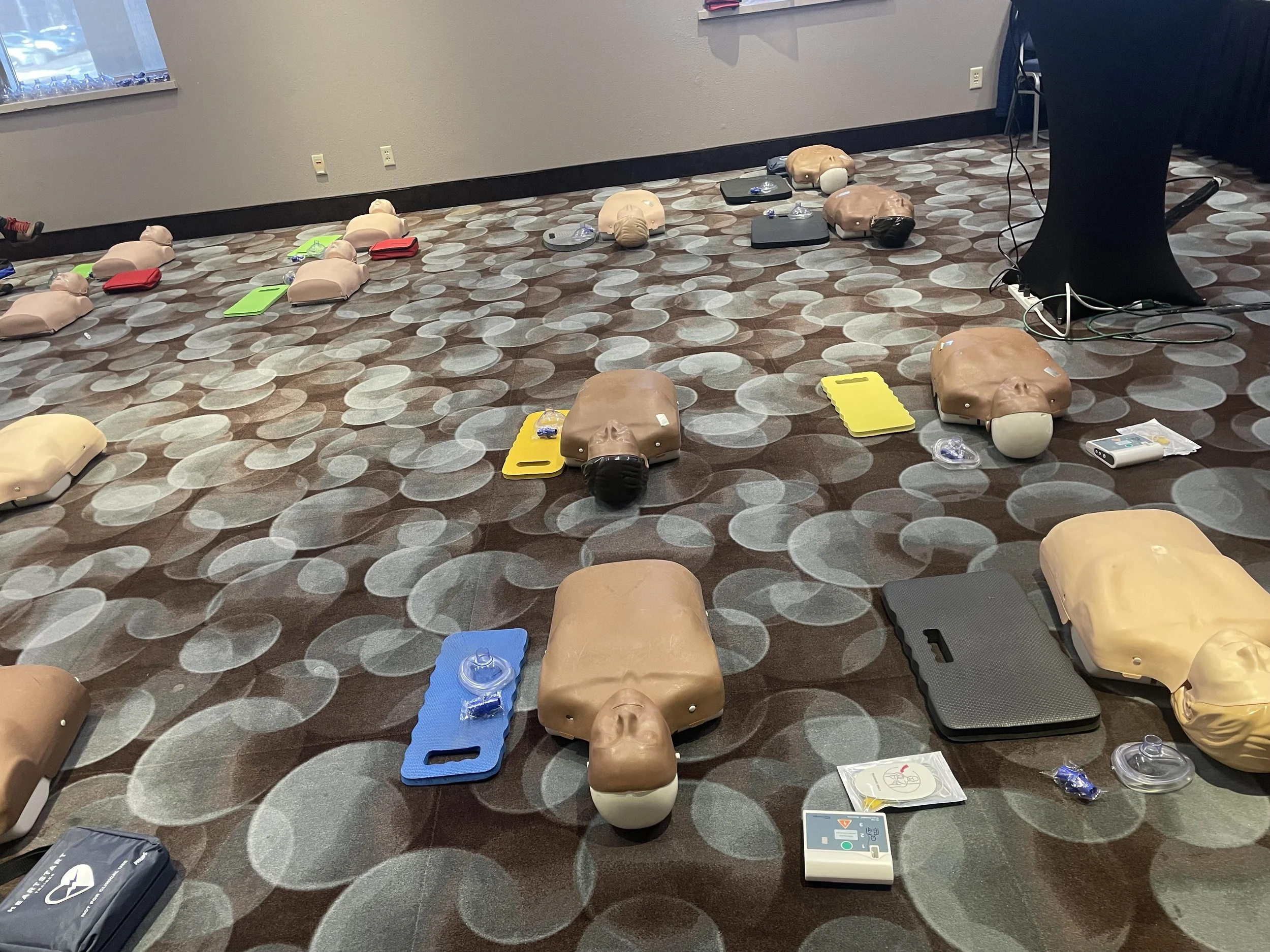 A room set up for CPR training with multiple practice mannequins, some with heads tilted back, placed on various colored mats on patterned carpet. There are ventilation masks, instructional handouts, and a defibrillator trainer on the floor.