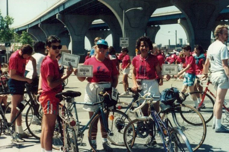 Group of people with bicycles gathered outdoors under an overpass, some holding certificates, during a cycling event in the 1980s.