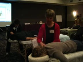 A woman in a red jacket demonstrates a massage technique on a person's back at a wellness event, with other attendees and a presentation screen in the background.