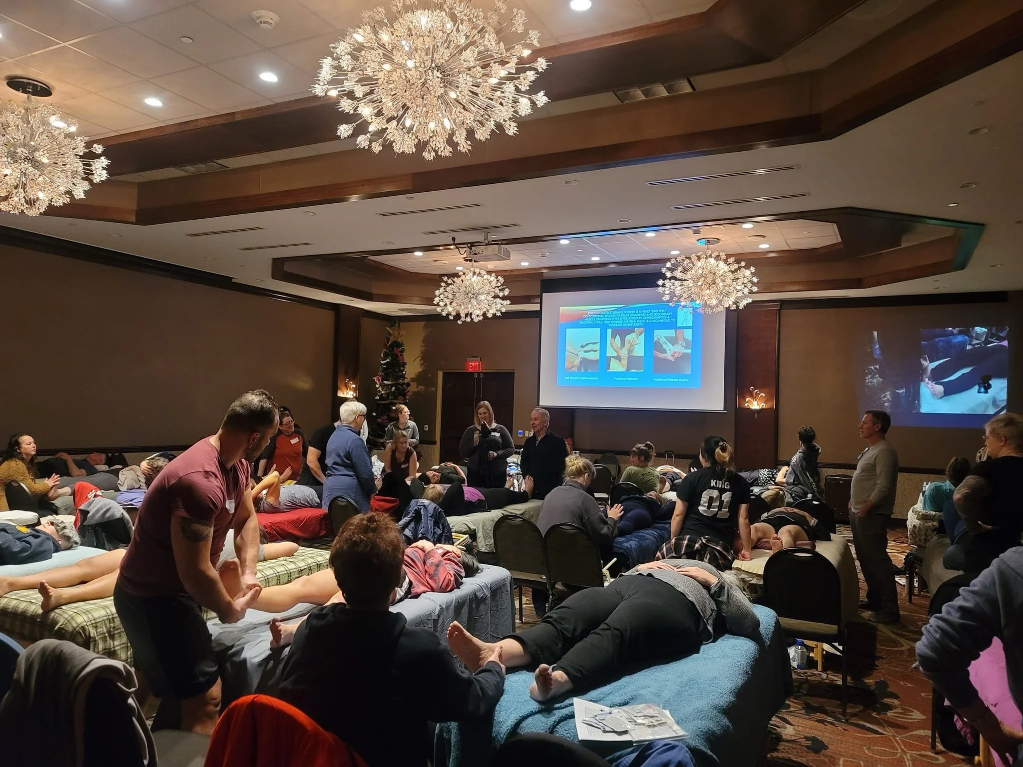 A large conference room filled with people. Some are lying on tables or mats, receiving massages. Others are sitting and watching a presentation on a large screen at the front of the room. Chandeliers hang from the ceiling, and there is a Christmas t