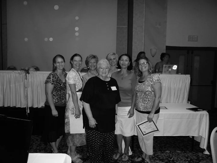 Six women standing together in a conference room, one holding a certificate, with a table and a laptop in the background.