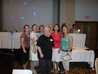 Group of seven women standing together in a conference room, some holding papers or folders, with a table and curtain in the background.