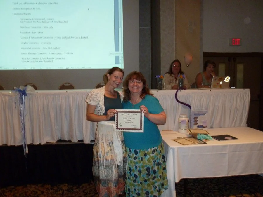 Two women smiling and holding a certificate at an award ceremony, seated at a table with a projector, in a conference room with two women seated at a table in the background.