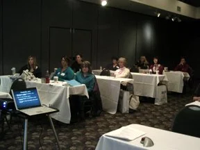 People seated at tables in a dark conference room, attending a presentation.