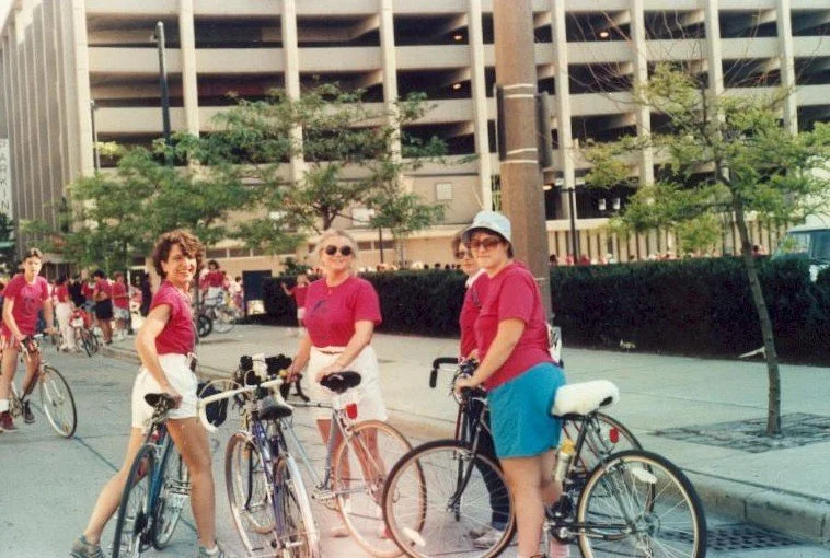 Three women with bicycles standing on a city street with a parking garage and trees in the background.