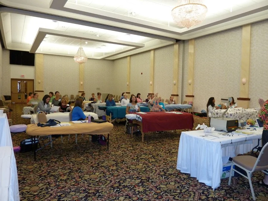 People participating in a massage therapy class or workshop in a large conference room, with massage tables, some students being massaged, and educational materials on a table.