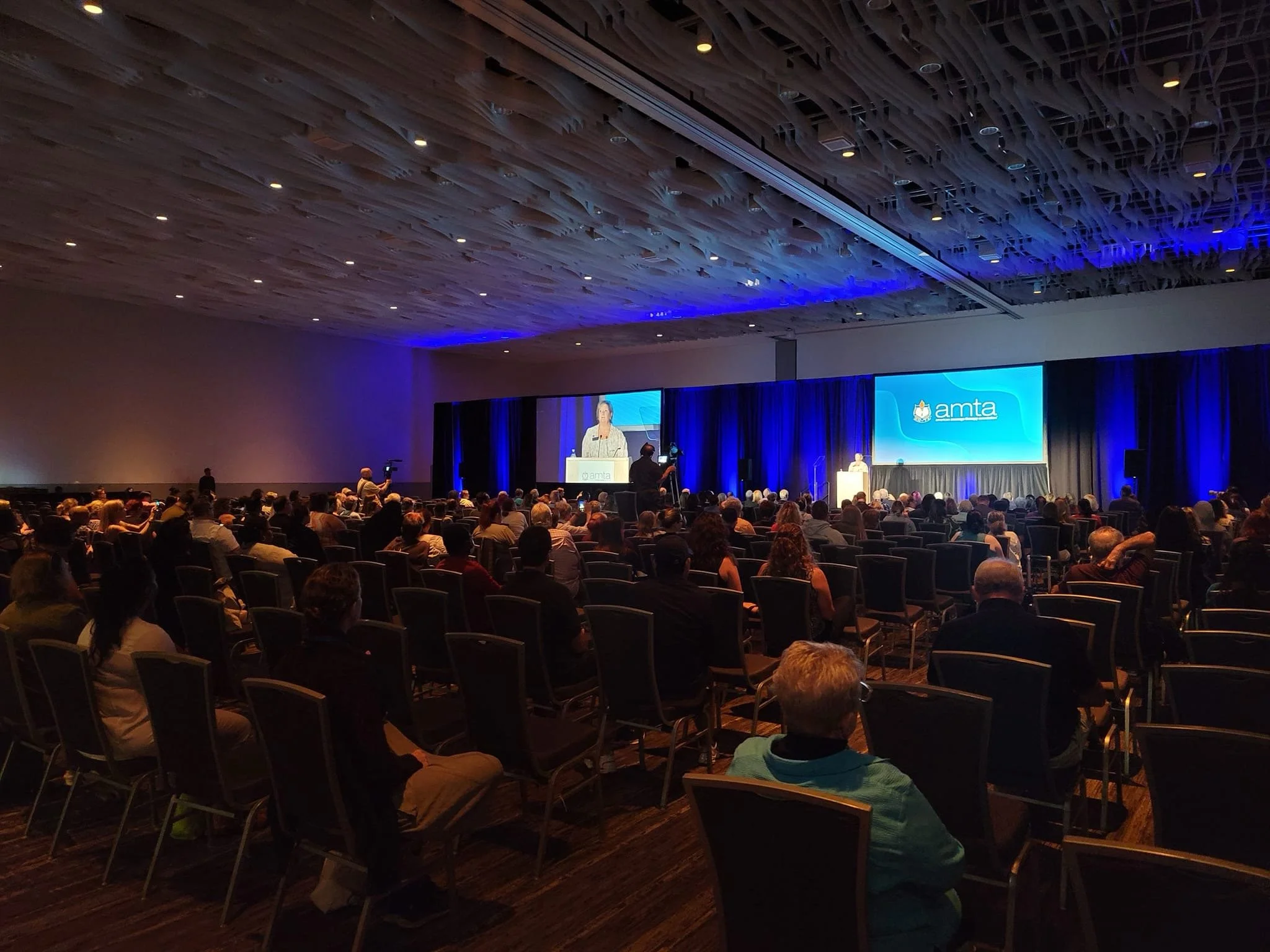 Conference attendees sitting in rows facing a stage with a large screen displaying the "amta" logo, in a dimly lit room with a dark wood ceiling.