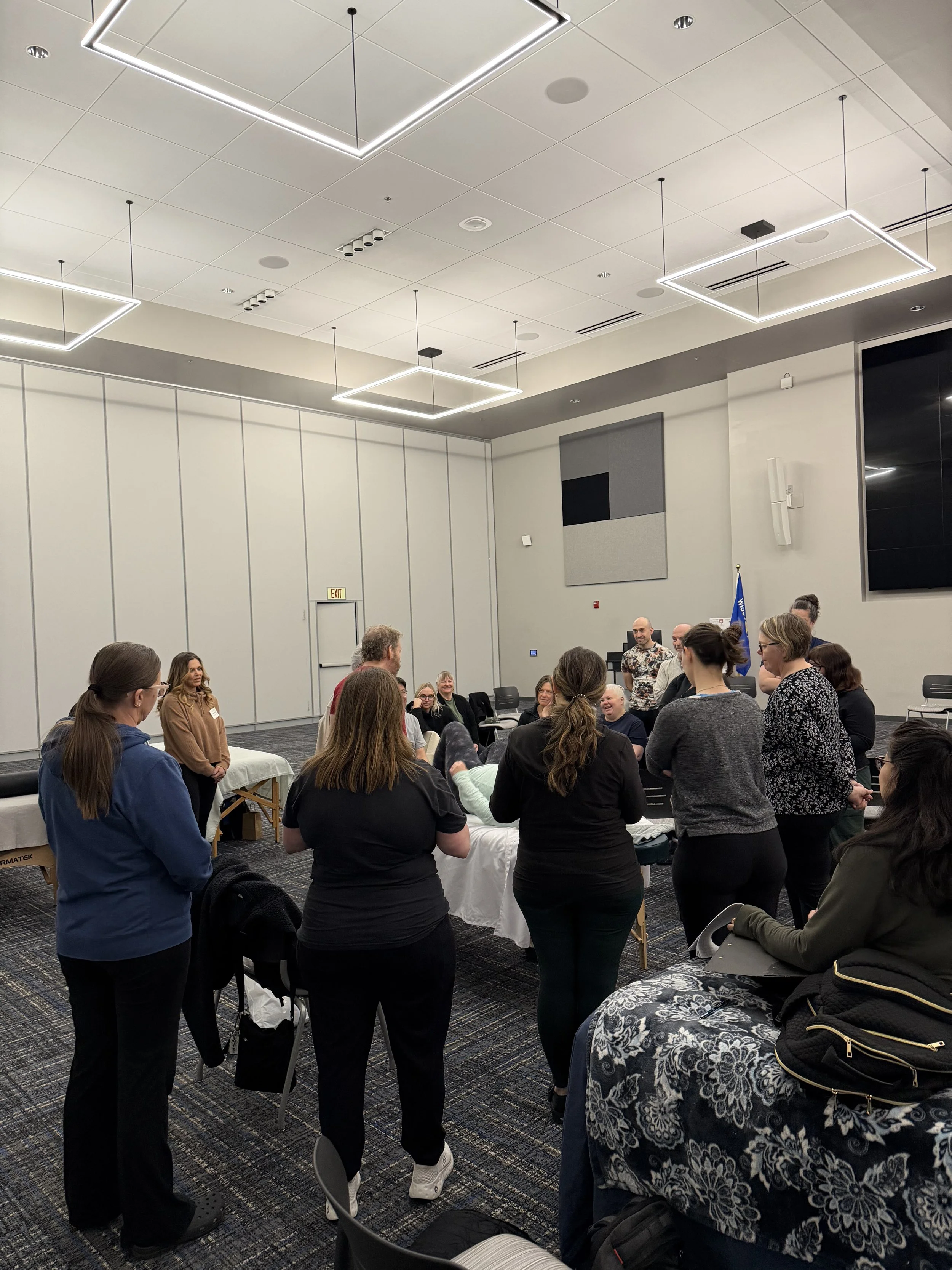 People gathered in a conference room, some seated and some standing, engaged in discussion or listening. The room has a modern ceiling with square lighting fixtures, gray walls, and a large screen or black panel on one wall. A group appears to be par