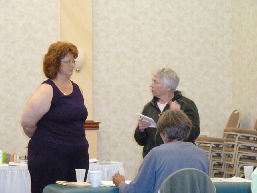 Three women in a conference room engaged in conversation, with one woman standing and two seated, one holding a notebook.