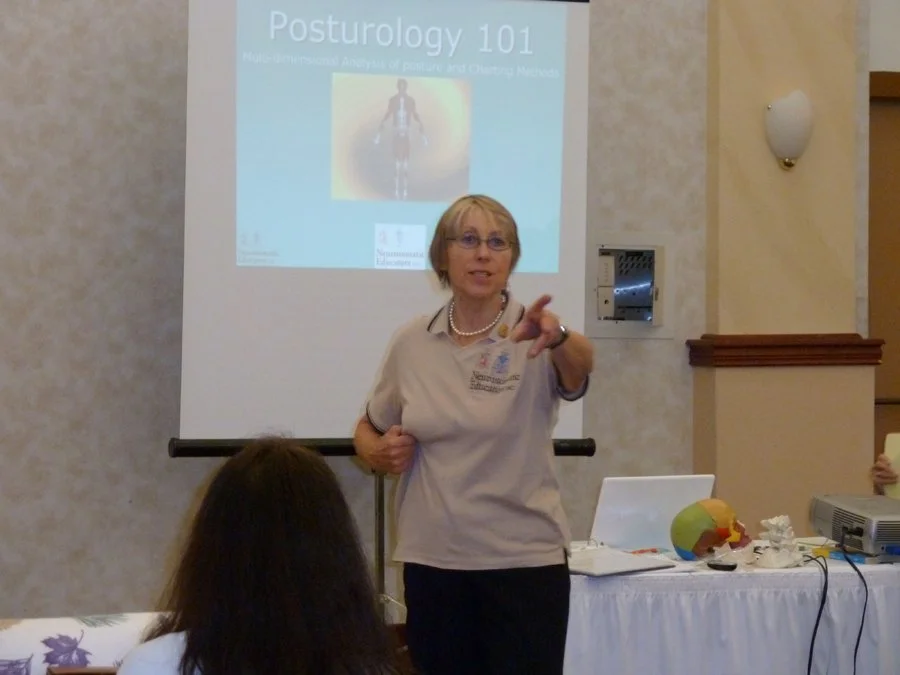 A woman giving a presentation about Posturology 101 in a conference room, pointing towards the audience, with a projection screen behind her displaying the title and an image of a human body.