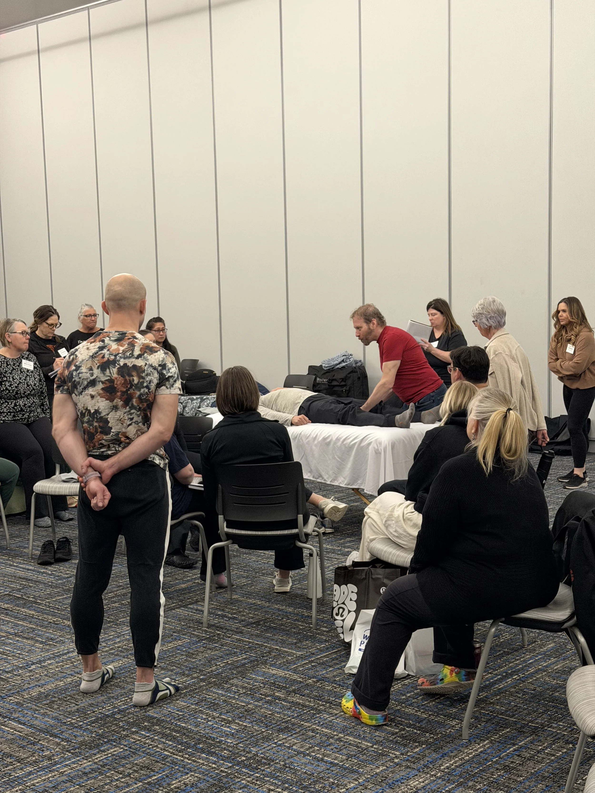 A group of people participating in a massage or therapy workshop, with some standing and some lying on tables, in a room with high beige walls and a patterned carpet.