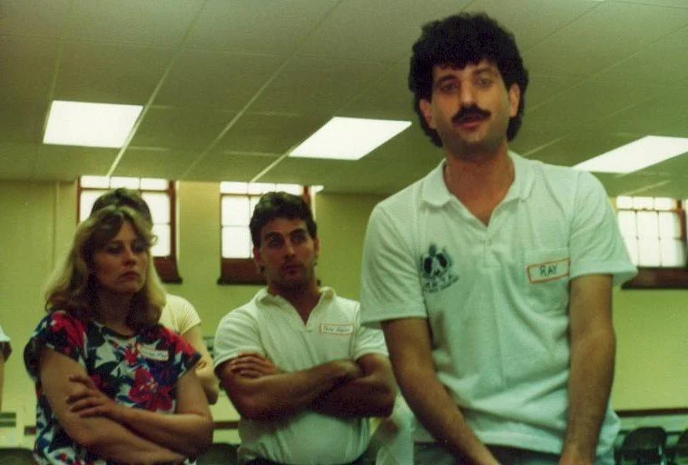 Three individuals in a room with green walls and large window with blinds. Two men and one woman are visible, with the man on the right wearing a white polo shirt with a name tag. The woman and the other man are in the background, with the woman cros