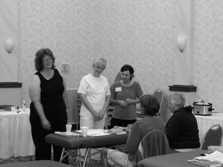 Four women in a meeting or workshop setting, with three women standing near a table and two women sitting, in a room with a high ceiling and wall-mounted lights.