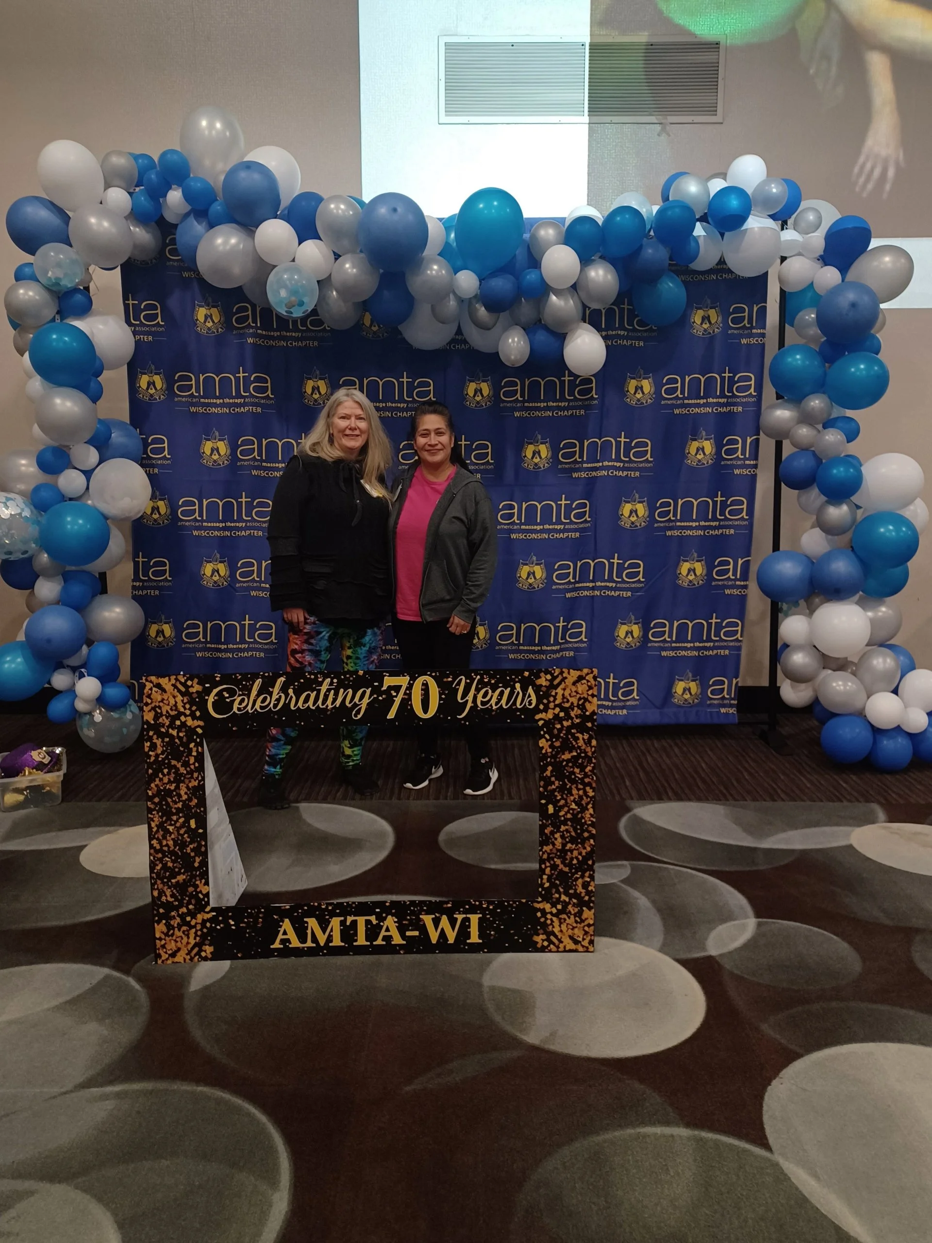 Two women standing in front of a blue backdrop with the logo and text of the American Massage Therapy Association Wisconsin Chapter. The backdrop is decorated with blue, white, and silver balloons arranged in an arch. There is a sign in front of them