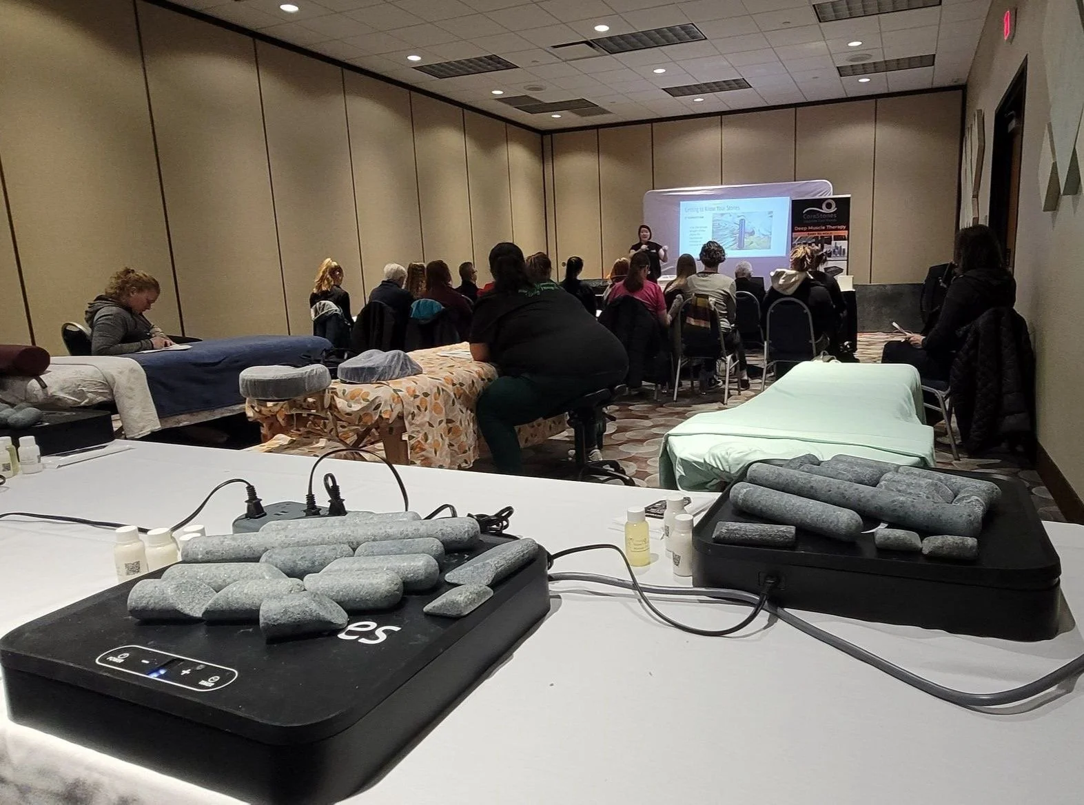 A group of people attending a presentation in a conference room. The image shows the audience seated while a speaker presents at the front. In the foreground, a table with a device and small bottles of lotion or oil.