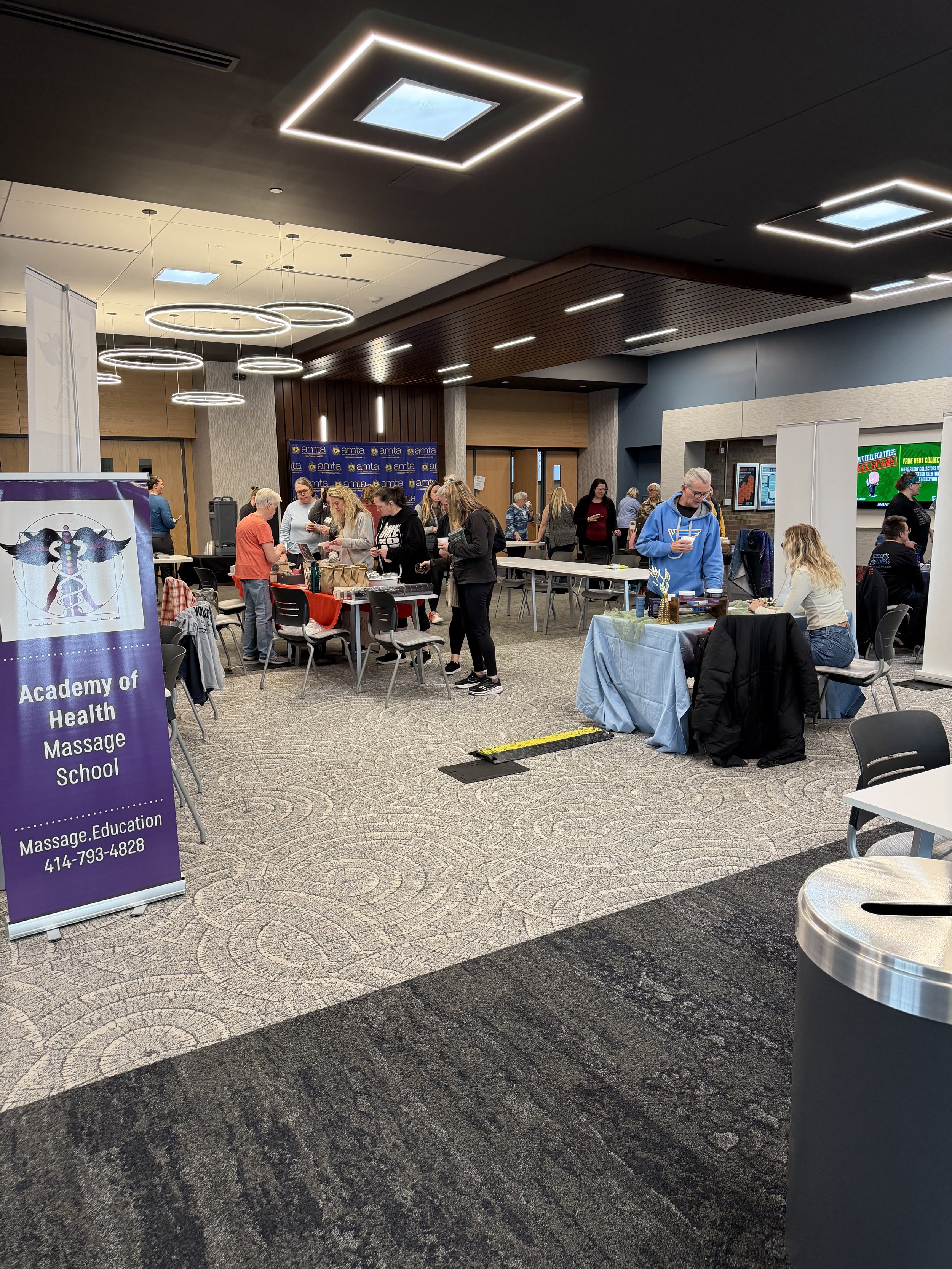 At a conference or event, people are gathered around tables with vendor booths and displays. There is a sign for Academy of Health Massage School, and attendees are engaging with different booths and talking. The room has modern lighting and a mix of