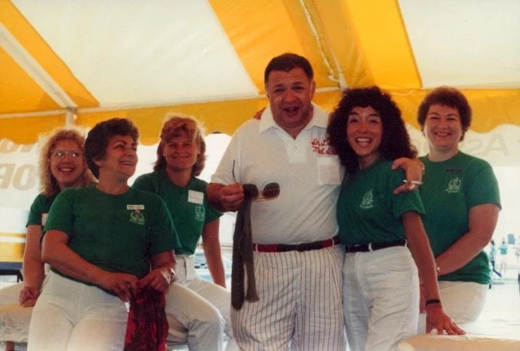 Group of five women and one man posing inside a yellow and white tent, smiling and enjoying themselves. The man is holding sunglasses and a scarf.