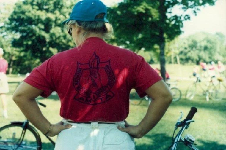 Back view of a woman in a red T-shirt with an emblem, white shorts, and a blue cap, standing outdoors with bicycles and trees in the background.