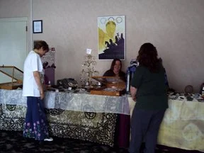 Three women at a banquet table with food and decorative items, one woman standing, two sitting behind the table, in an indoor setting with beige walls and a colorful painting on the wall.