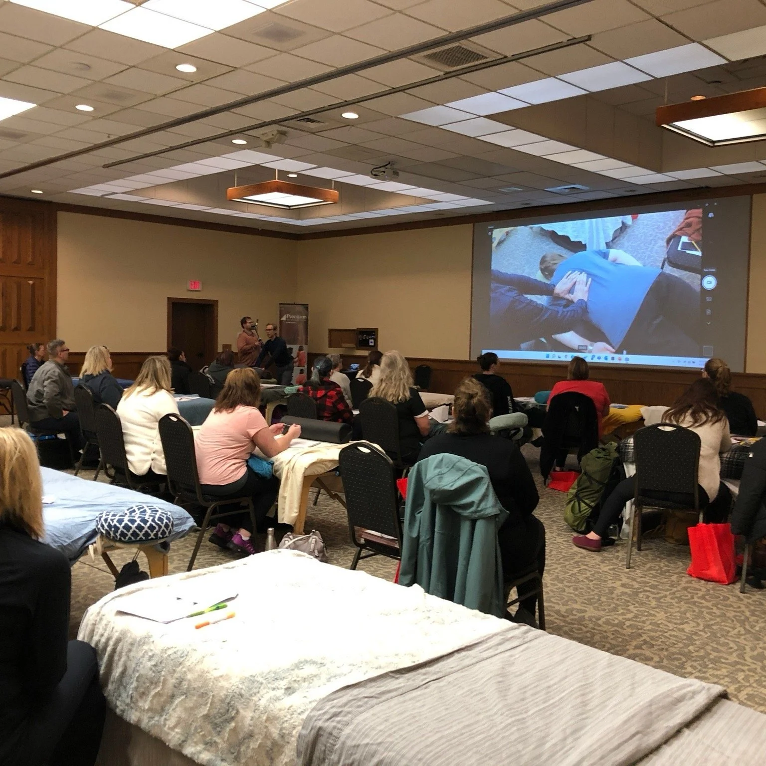 People attending a conference or workshop in a large room, some taking notes, while a presentation is projected on a large screen at the front of the room.