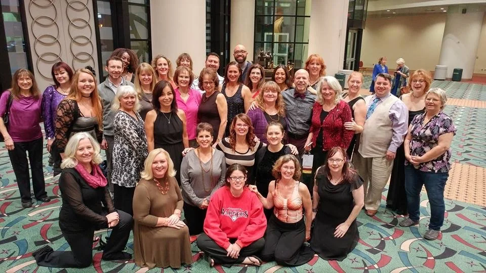 Group of about 30 people, mostly women, posing for a photo in a large indoor lobby with modern decor and a patterned carpet.