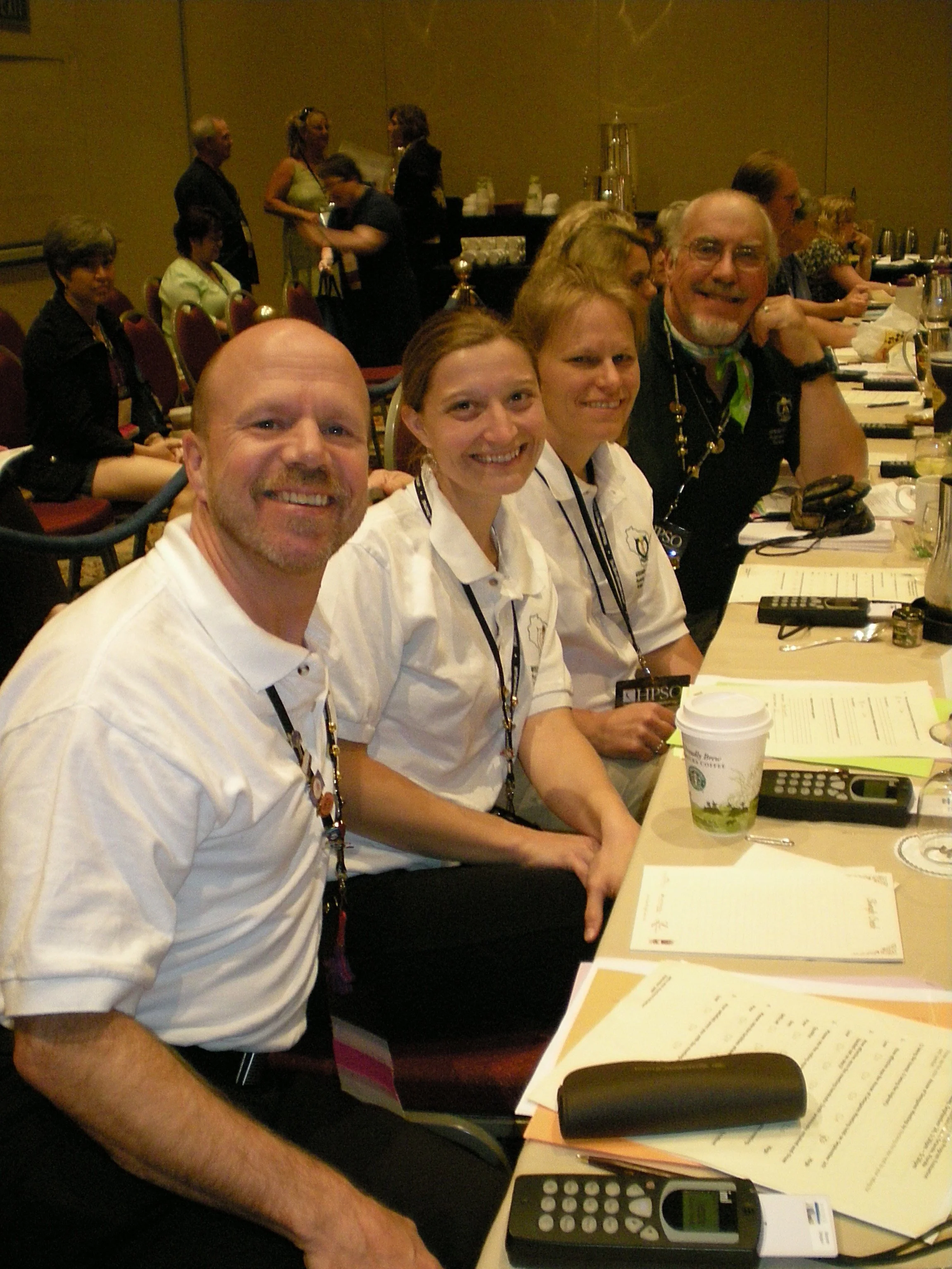 Four people sitting at a conference table with papers, drinks, and electronic devices, smiling at the camera, while others stand and converse in the background.