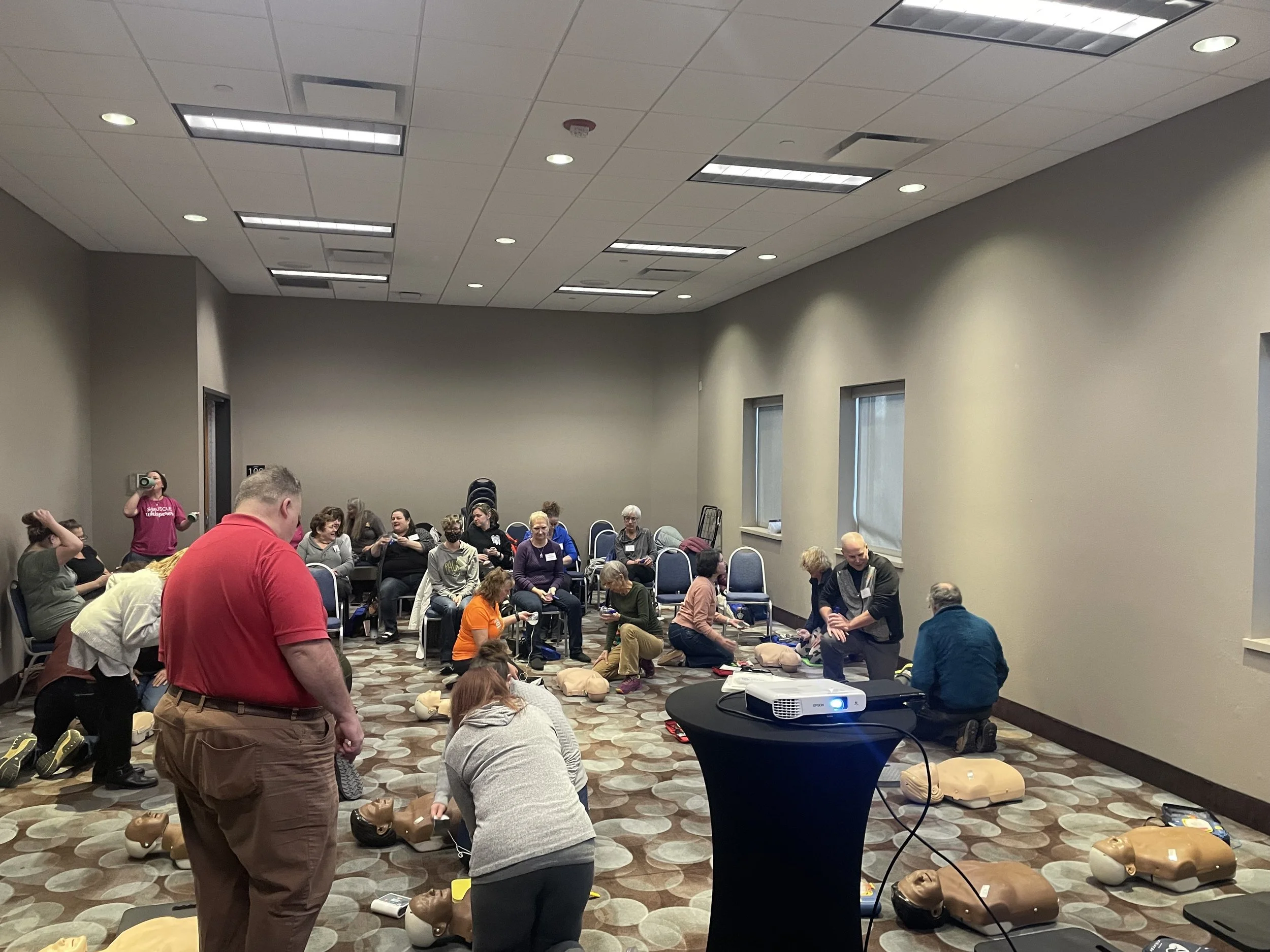 People participating in a CPR training session in a conference room, practicing on mannequins, with a CPR dummy on the floor and a projector on a table.