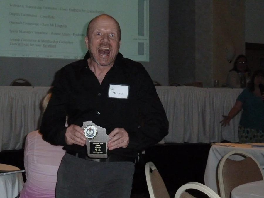 A man with a surprised or excited expression holding an award plaque while standing at a conference or event, with a presentation slide projected in the background and other people in the room.