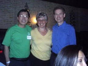 Three people standing together, smiling indoors against a brick wall. The person on the left is wearing a green shirt, the woman in the middle is in a yellow top, and the man on the right is in a blue shirt.