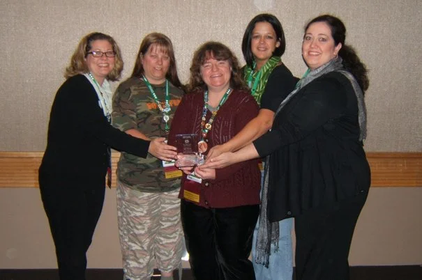Five women standing together indoors, holding a plaque or award, smiling at the camera.