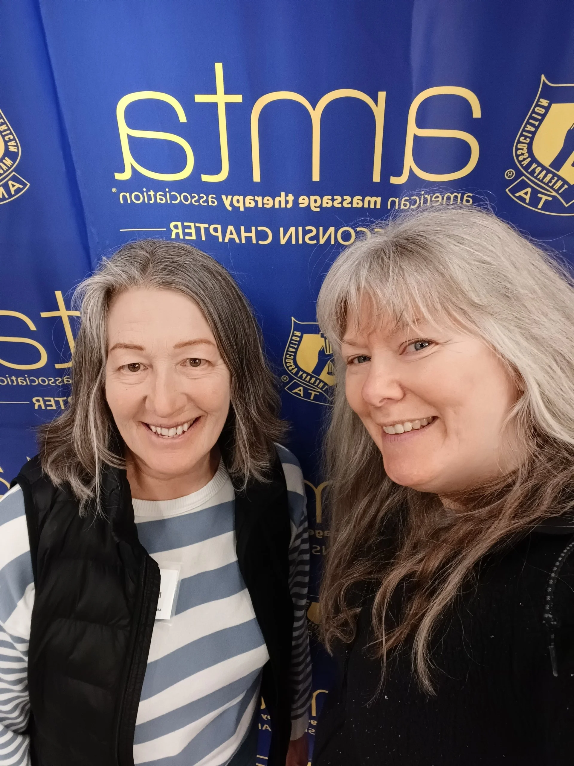 Two women smiling for a photo in front of a blue background with yellow text and symbols, likely at an event or conference.
