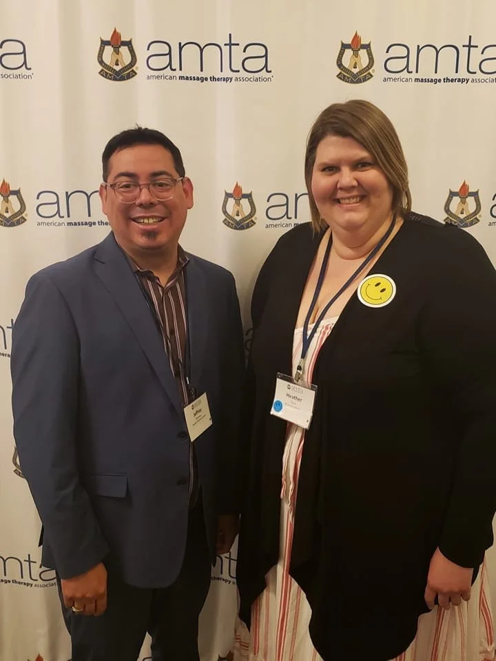 Two people, a man and a woman, standing together at an event with a backdrop displaying the American Massage Therapy Association (AMTA) logo. They are smiling, and the woman has a yellow smiley face pin on her jacket.