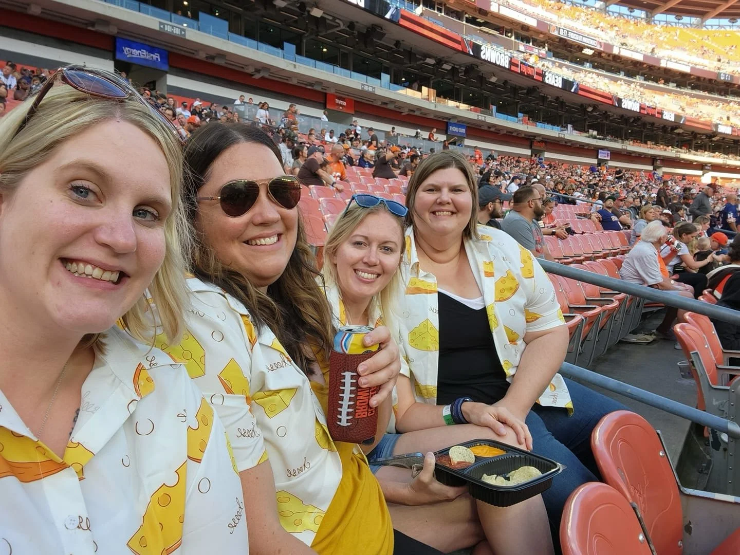 Four women sitting in stadium seats at a sports event, dressed in matching food-themed shirts with cheese and pizza graphics, smiling at the camera, with more spectators in the background.