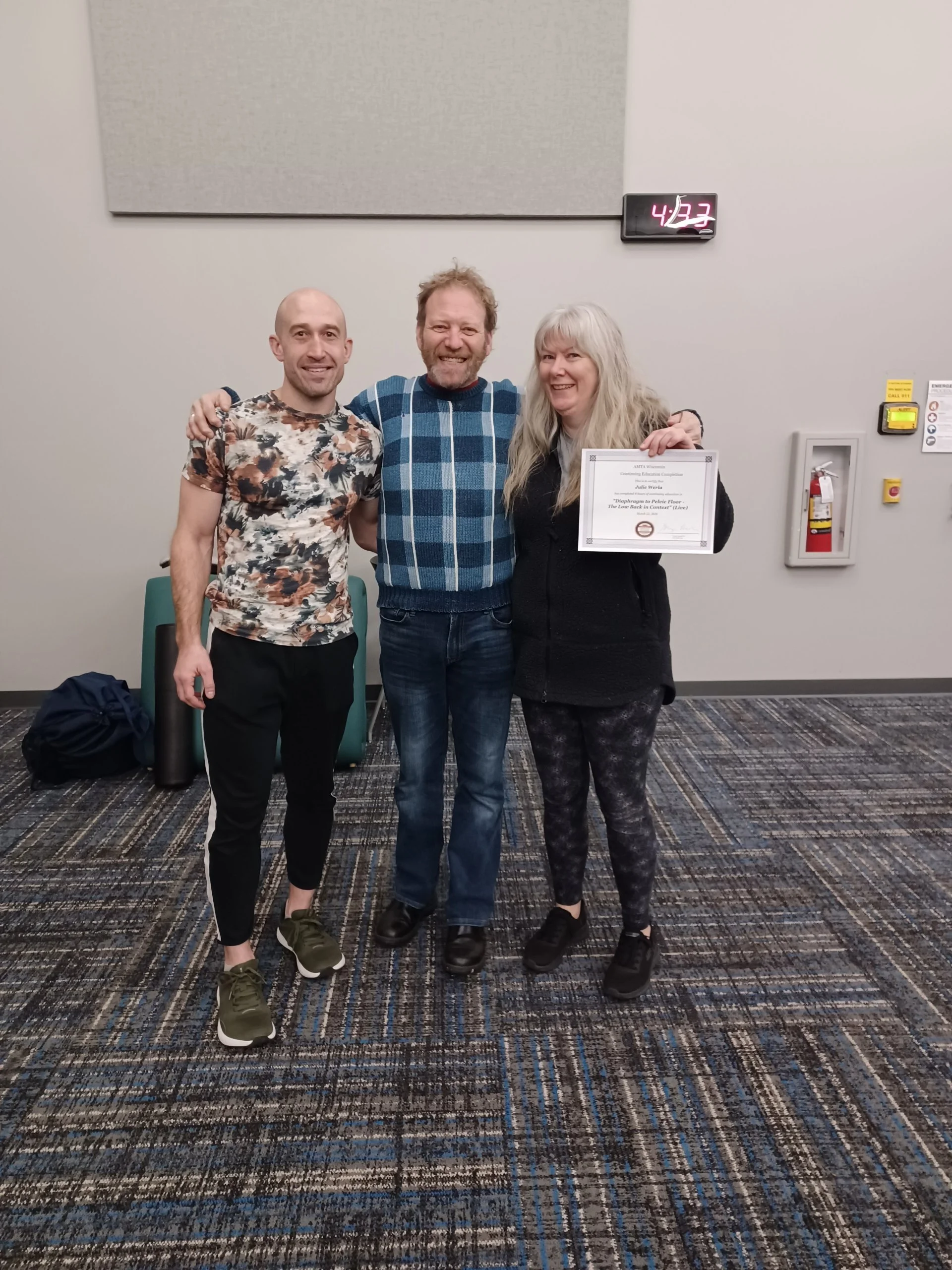 Three people smiling, standing close together indoors, with a certificate held by the woman on the right, colorful carpet, and a wall clock showing 4:23.