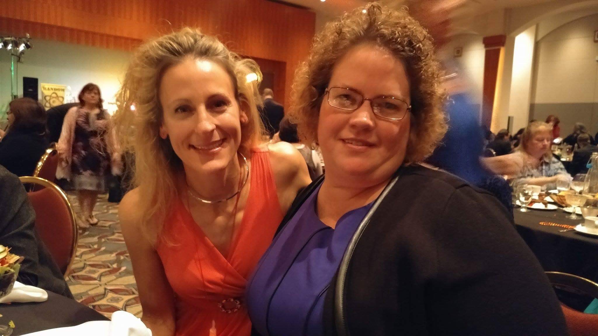 Two women smiling at a formal event or dinner, sitting close together at a table with other guests in the background.
