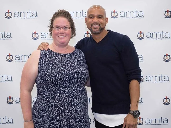 A smiling woman with curly hair and glasses standing next to a smiling man with a beard, wearing a black sweater. They are posed in front of a backdrop with the 'AMTA' logo repeated multiple times.