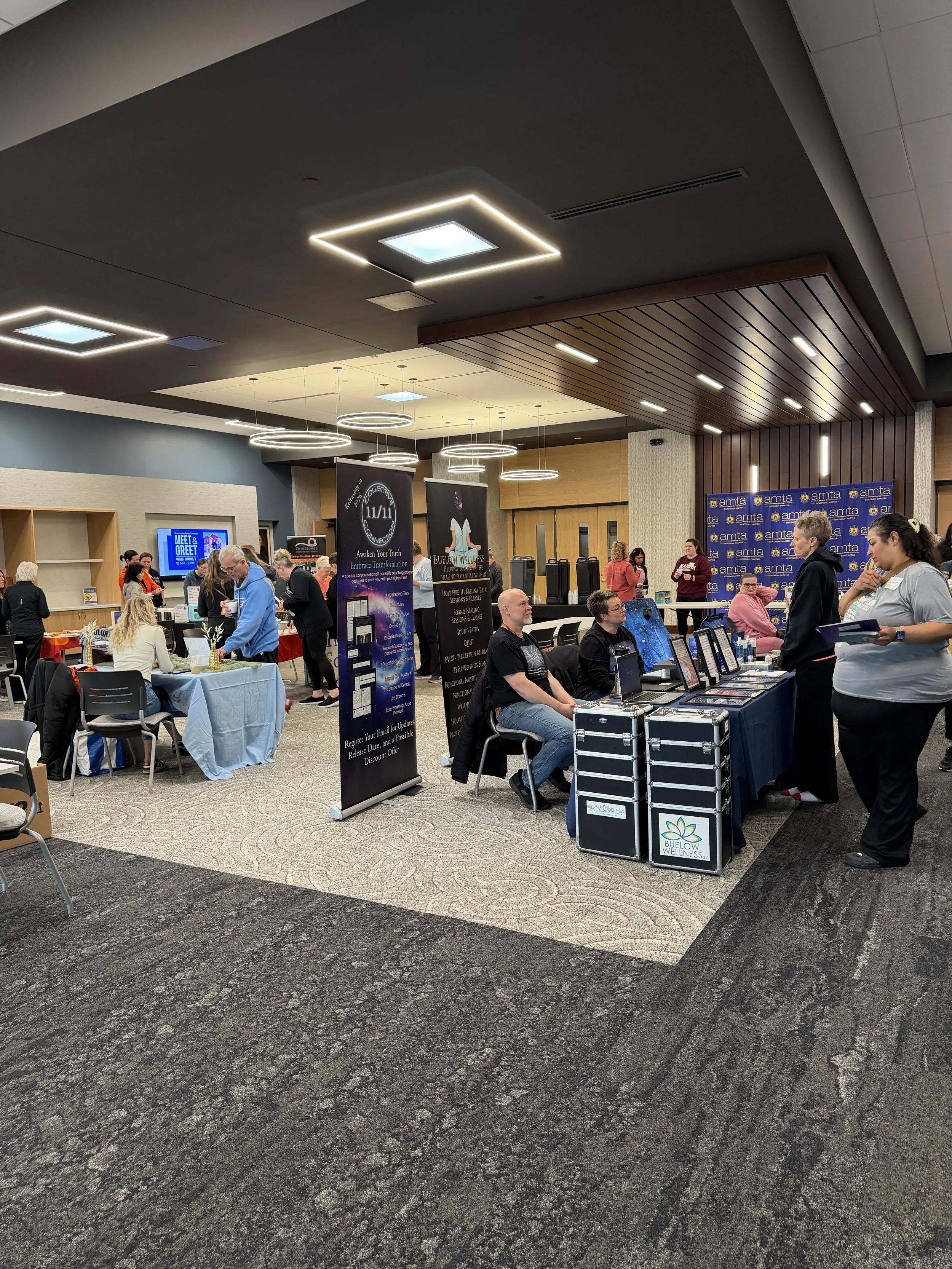 A busy indoor event with several booths and people engaging in conversations. There are tables with informational displays, a 'Meet & Greet' screen, and banners for wellness and connectivity. The ceiling features modern lighting fixtures, and the floor is carpeted with a textured pattern.