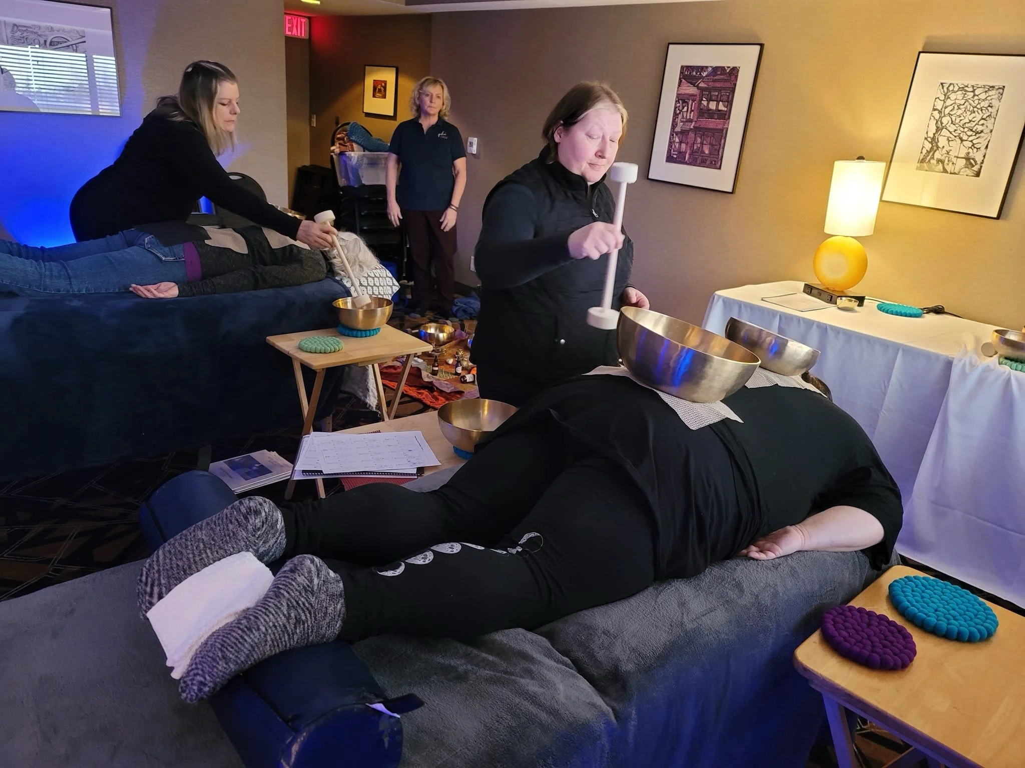 People participating in a sound healing session, lying on couches with their eyes closed. Two women are playing crystal singing bowls, and a woman in the background observes. The room is decorated with artwork and soft lighting.