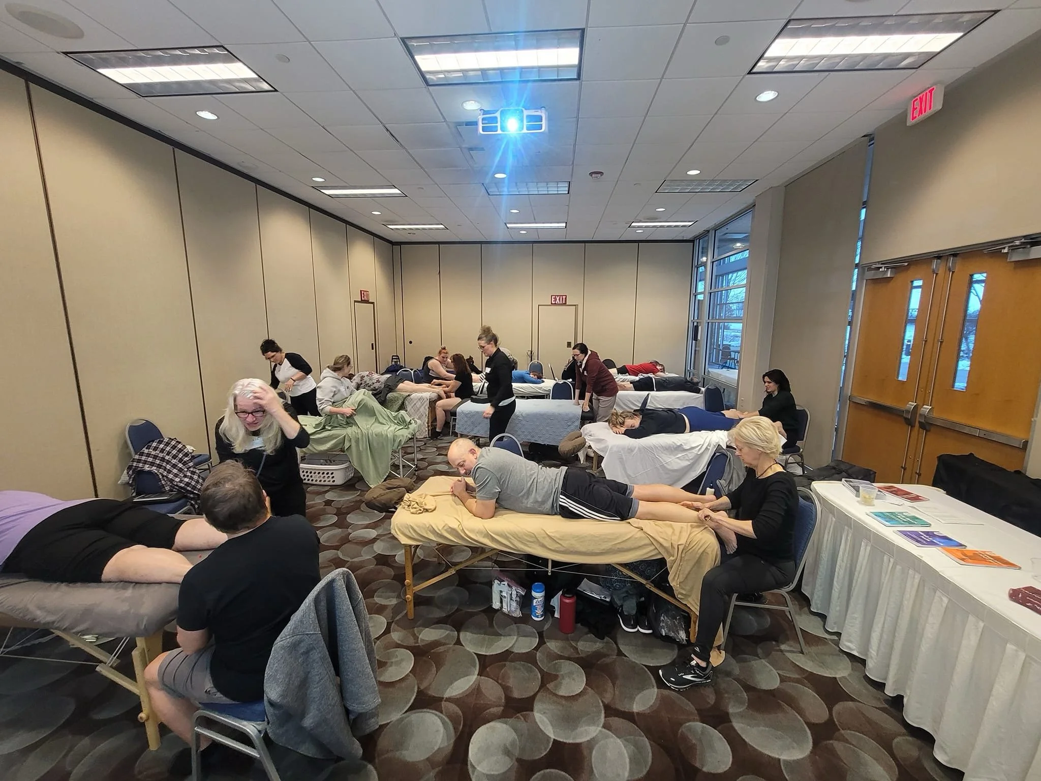 People lying on massage tables receiving massages in a conference room with beige walls, large windows, and a patterned carpet.