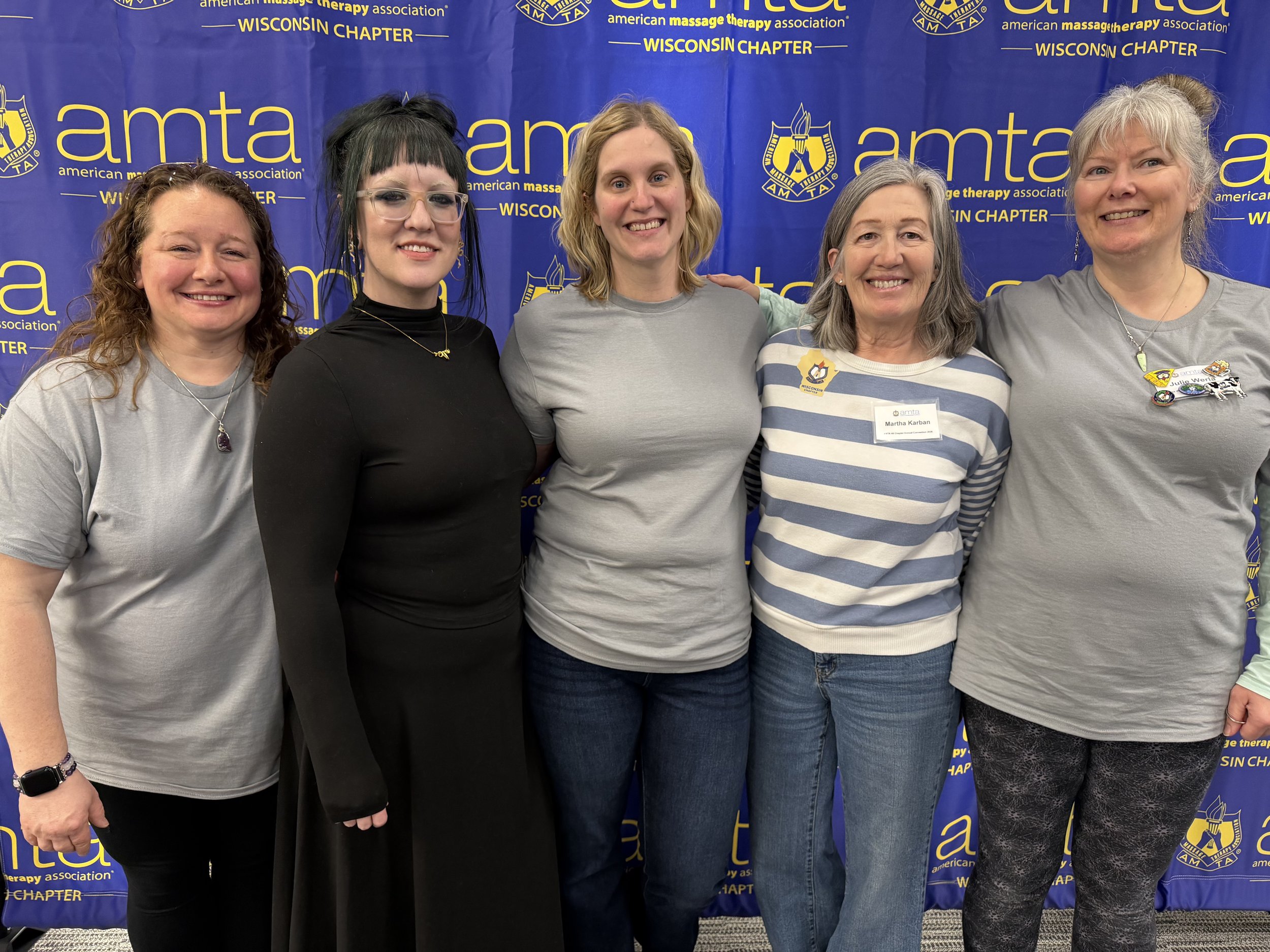 A group of five women standing close together, smiling, in front of a blue backdrop with yellow text and logos for the American Massage Therapy Association Wisconsin Chapter.