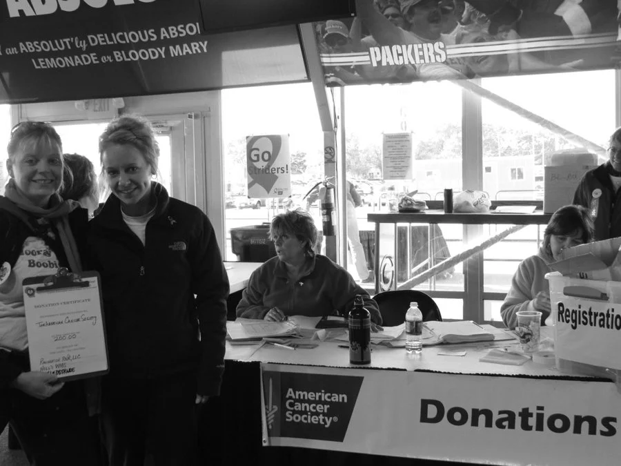 Two women smiling and holding a check at a charity event for the American Cancer Society, with registration and donation tables in the background.