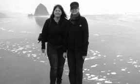 Two people standing on a beach with rocks and water in the background.