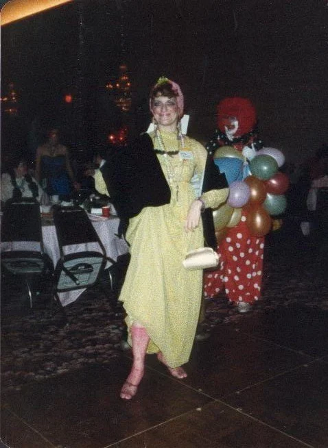 Woman in a yellow dress and pink headscarf standing in a dimly lit event space, with a clown holding balloons in the background.